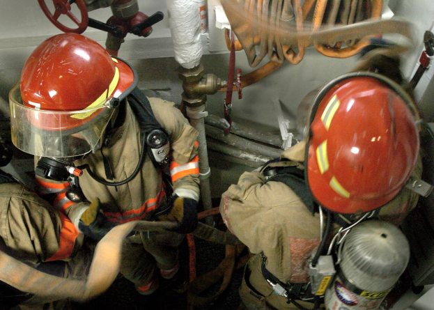 Feuerwehrleute in Helmen und Schutzausrüstung arbeiten an einem Feuerhydranten mit Rohren, Ventilen und einer Wand im Hintergrund; Text ist unten sichtbar.