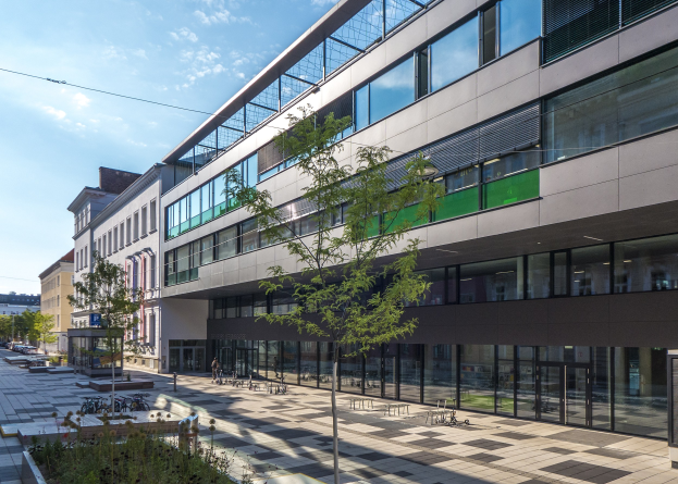 Großes modernes Gebäude mit zahlreichen Fenstern, umgeben von Bäumen und Grünfläche, mit parkenden Fahrzeugen und Fußgängern, unter einem bewölkten Himmel, identifiziert als neue deutsche Technologie-Institut-Zentrale in Berlin.