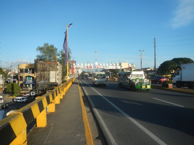 Vollgestopfte Stadtstraße mit mehreren Fahrzeugen, einer Mitteltrennung, einem Fahnenmast mit Fahne, Gebäuden, Bäumen, Strommasten mit Drähten und einem bewölkten Himmel.