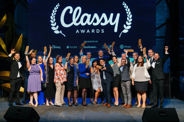 Gruppe von Menschen auf einer Bühne, die Auszeichnungen halten, mit Lautsprechern unten und einem Bildschirm im Hintergrund mit "Classy Awards 2019"