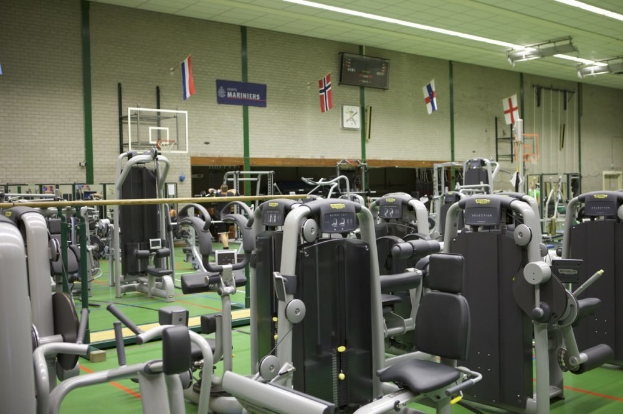 Ein Innenraum eines Fitnessstudios mit Sportgeräten, einer Wand im Hintergrund, Deckenleuchten und mehreren Menschen.