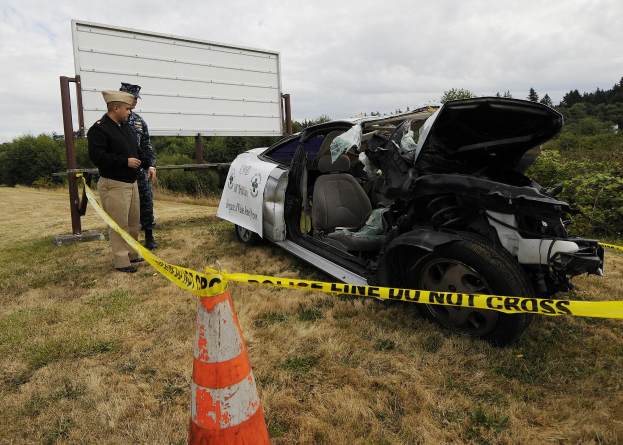 Ein Polizist in Uniform steht neben einem schwer beschädigten Auto am Straßenrand mit eingedellter Haube und zersplitterten Fenstern neben einem Verkehrskegel und einer markierten Stange.