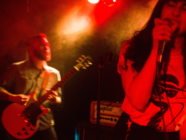 Eine Frau singt in ein Mikrofon auf der Bühne, wöhrend ein Mann im Hintergrund Gitarre spielt, mit Deckenleuchten, die die lebendige Szenerie beleuchten.