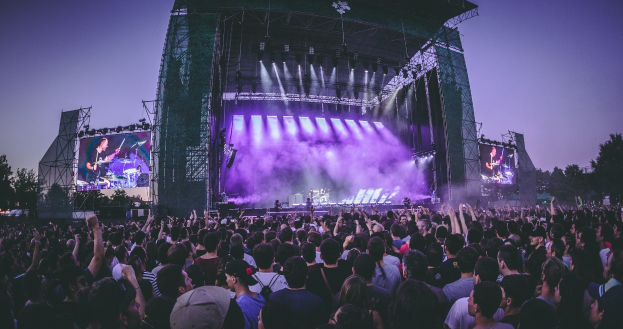 Eine große Menschenmenge bei einem Musikfestival schaut auf eine hell erleuchtete Bühne mit Instrumenten und Bildschirmen, die vor einem Gebäude, Bäumen und einem klaren blauen Himmel liegt.