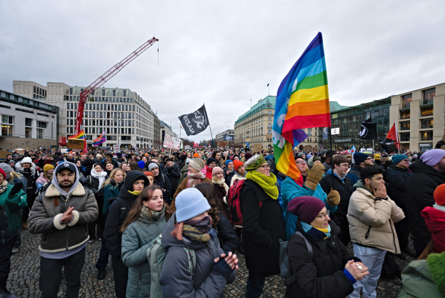 Große Gruppe von Menschen mit LGBTQ+-Rechten-Schildern vor einem Gebäude mit einem Kran und einem bewölkten Himmel.