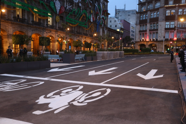 Stadtstraße mit einem ausgewiesenen Fahrradweg, umgeben von Gebäuden, Straßenmöblierung, Fußgängern, Grünflächen und einem klaren blauen Himmel.