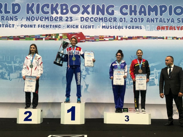 Fünf Menschen auf einem Podium mit Trophäen und Zertifikaten und einem "World Kickboxing Championship Antalya 2019"-Plakat im Hintergrund.