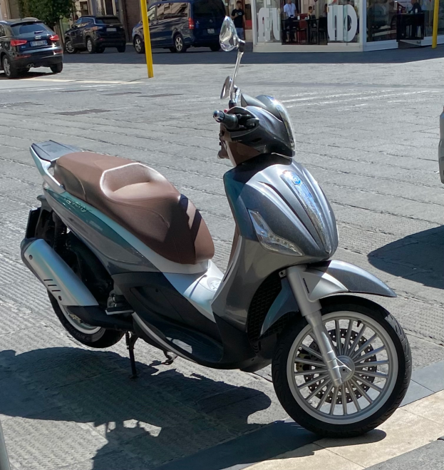 Ein Motorroller am Straßenrand geparkt mit einem Helm darauf, mit Gebäuden, Bäumen, Polen, Menschen, Stühlen, Tischen und anderen Gegenständen im Hintergrund.