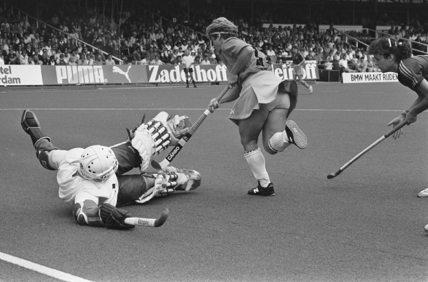 Schwarzes und weißes Bild von Frauen, die Feldhockey in einem Stadion mit Zuschauern in den Rängen spielen, Helme tragen und Stöcke halten, umgeben von Tafeln mit Text.