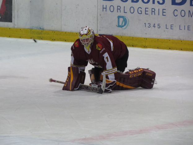 Eishockey-Spieler in rot-gelber Uniform, der einen Schuss auf dem Eis abwehrt, trägt Helm, Handschuhe und Knieprotektoren und hält einen Eishockey-Schläger, mit einer Wand und Text im Hintergrund.