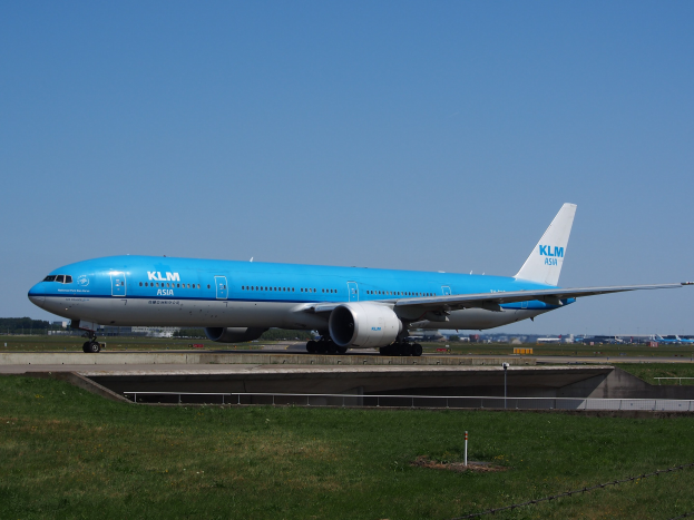 KLM Boeing 777-300ER auf der Rollbahn am Amsterdam Airport, umgeben von Gras und einem Zaun mit Pfählen, mit Gebäuden, Bäumen und einem klaren blauen Himmel im Hintergrund.