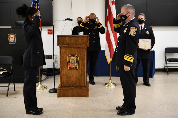 Gruppe uniformierter Personen in Masken vor einem Podium mit Mikrofon, umgeben von Fahnen, Stühlen und Tischen während einer Vereidigung für einen neuen Feuerwehrchef.