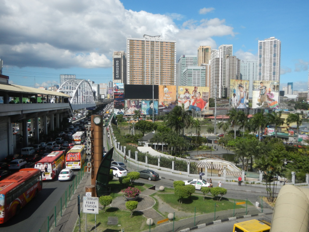 Eine belebte Stadtstraße mit Bussen, Autos und anderen Fahrzeugen, gesäumt von Bäumen, Pflanzen, Gras, Pfählen, Plakaten, Gerüsten und einer Brücke im Hintergrund, mit Gebäuden und einem bewölkten Himmel.