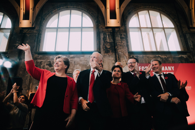 Eine Gruppe von Menschen, Mitglieder der Labour Party, klatschen feierlich vor einer Menge, mit einem Banner, Glasfenstern und beleuchteten Lichtern im Hintergrund.