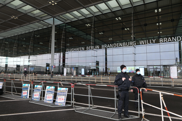 Zwei Polizisten in schwarzen Uniformen stehen vor einem Glasgebäude mit Text, mit Absperrungen und Schildern im Vordergrund.