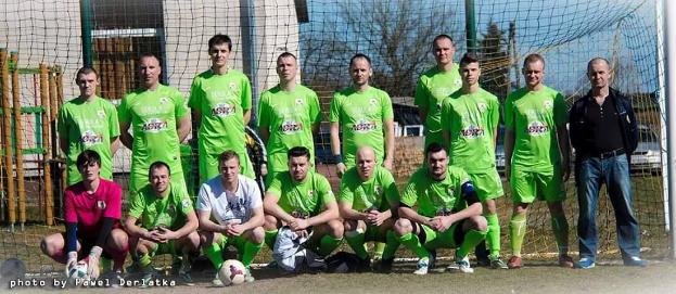 Eine Gruppe von Männern in grünen T-Shirts steht auf einem Fußballfeld, einige hocken, mit einem Ball und einem Torpfosten zu sehen; der Hintergrund enthält Bäume, Gebäude und Fahrzeuge.