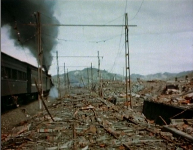 Ein Zug fährt durch ein beschädigtes Gebiet mit Rauch, der aus ihm aufsteigt, umgeben von Strommasten und -leitungen, Hügel im Hintergrund unter einem klaren Himmel und verstreute Trümmer auf dem Boden.