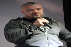 Ein Mann in schwarzer Kapuzenjacke und schwarzem Anzug mit weißem Hemd, der selbstbewusst mit zurückgekämmtem Haar steht und ein Mikrofon vor einer Wand hält.