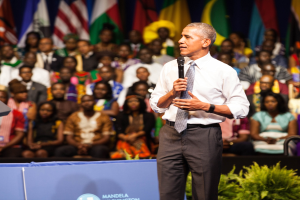 Präsident Obama spricht auf einer Wahlkampfveranstaltung vor einer Menge, hält ein Mikrofon, vor Flaggen und einer Tafel mit Text, mit einer Pflanze im Vordergrund.
