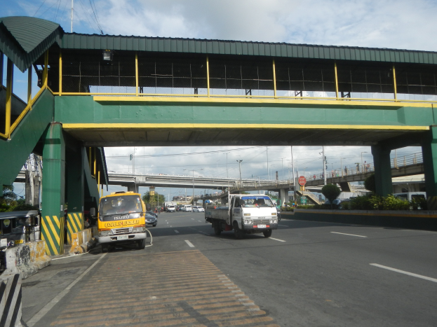 Eine Mautstation mit Fahrzeugen auf der Straße darunter, eine Brücke mit Geländern und Pfeilern, Laternenmasten, Schilder an Masten, Bäume und ein bewölkter Himmel im Hintergrund, mit einem Lastwagen, der neben der Brücke fährt.