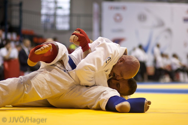 Zwei Judoka im Kampf, mit Zuschauern und Indoor-Elementen im Hintergrund.