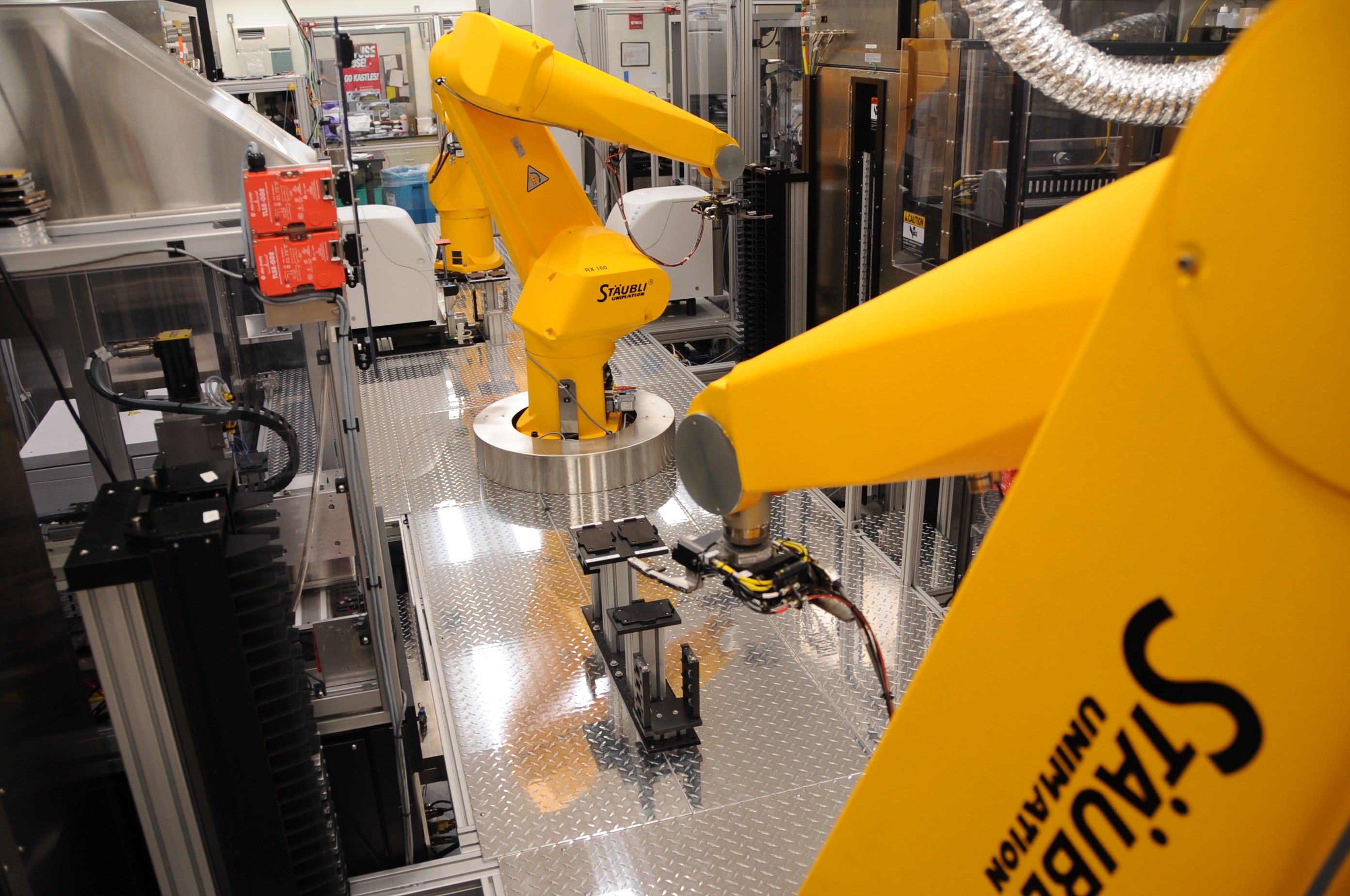 Ein gelber Industrieroboter bedient Maschinen in einer hell erleuchteten Fabrikumgebung mit Rohren und anderen Geräten im Hintergrund.