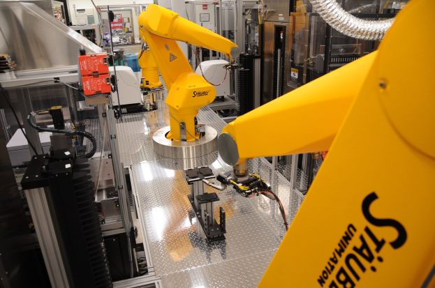 Ein gelber Industrieroboter bedient Maschinen in einer hell erleuchteten Fabrikumgebung mit Rohren und anderen Geräten im Hintergrund.