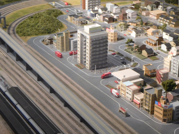 Ein detailliertes Miniaturmodell einer Stadt mit einem Zug auf Schienen, umgeben von Gebäuden, Bäumen, Straßen, Fahrzeugen, Strommasten, Gras und einem Wolkenkratzer.