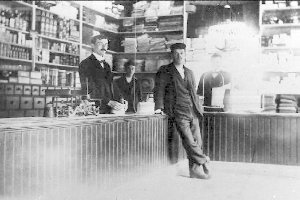 Drei Männer stehen an einem Tresen in einem Laden, umgeben von verschiedenen Gegenständen, mit einem Regal voller Bücher und anderen Gegenständen im Hintergrund, die scheinbar an einem Überfall beteiligt sind.