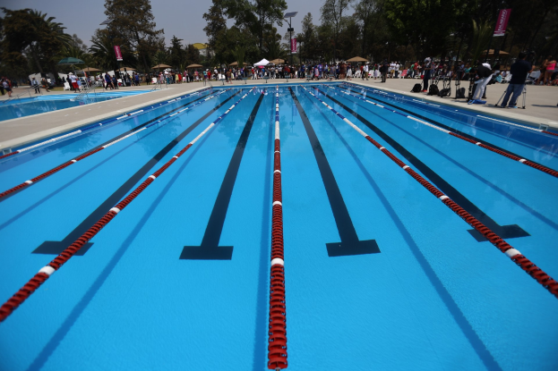 Großer Schwimmbad mit Bahnentrennung, umgeben von Menschen mit Taschen und Gegenständen, mit Zelten, Transparenten, Stangen, einem Schild und einem bewölkten Himmel im Hintergrund.