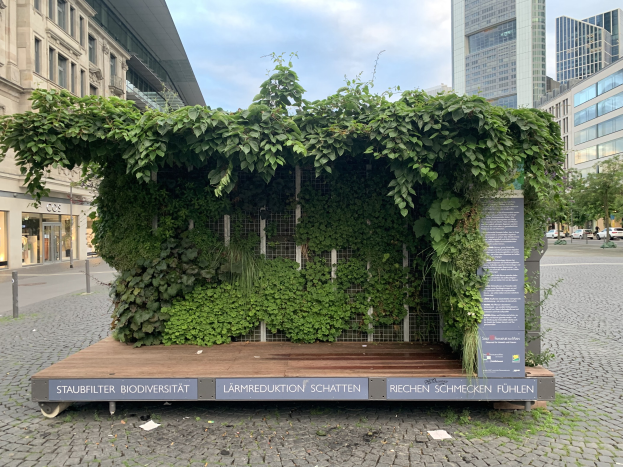 Ein grünes Gebäude mit Efeu an der Seite, umgeben von Bäumen, Fahrzeugen, Strommasten, Schildern und anderen Gebäuden unter einem bewölkten Himmel, Teil des nachhaltigen Staufbilter Biodiversität-Gebäudes in Berlin, Deutschland.