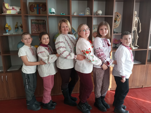 Eine Gruppe von Kindern in traditioneller ukrainischer Kleidung steht vor einem Regal, das mit Spielzeugen, einem Skelett und anderen Gegenständen gefüllt ist, was auf eine ukrainische Schulumgebung hinweist.