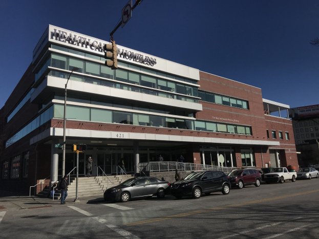 Großes Gebäude mit der Aufschrift "New York City Health Care Homeless Center" und parkenden Autos, Fußgängern, Straßenmöbeln und Bäumen unter einem klaren Himmel.