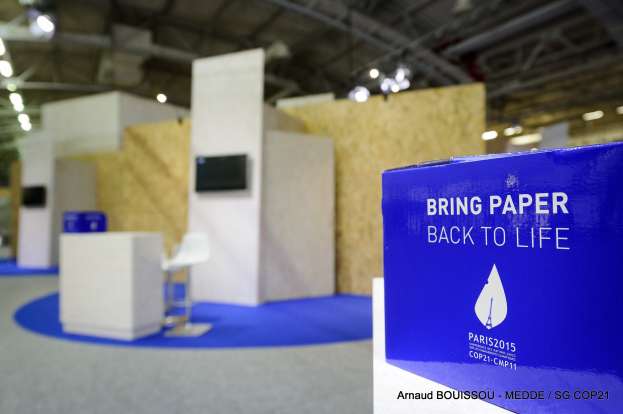 Messestand mit einem blauen und weißen "Bring Paper Back to Life"-Schild auf der rechten Seite, das einen blauen Stuhl, einen Tisch, einen an der Wand montierten Fernseher und eine Deckenbeleuchtung im Hintergrund zeigt.
