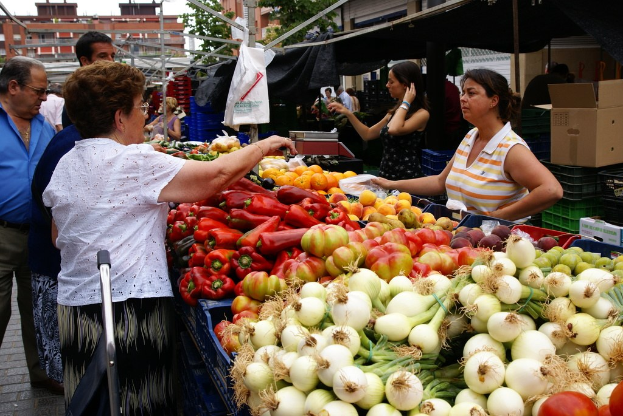 Gemüse in Plastikbehältern, Menschen in einer Gruppe, Gebäude, Bäume, eine Kartonschachtel und der Himmel.