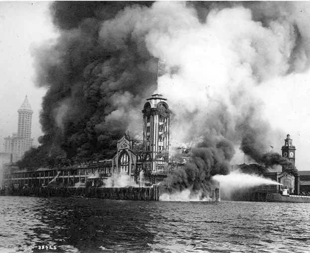 Schwarz-weiß-Foto eines großen, von Flammen umloderten Gebäudes mit aufsteigendem Rauch, einem Gewässer im Vordergrund und anderen Gebäuden sowie dem Himmel im Hintergrund.