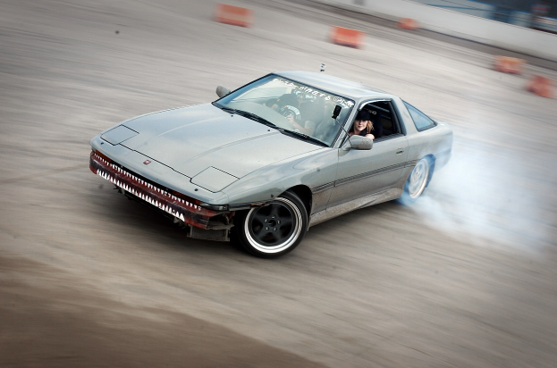 Eine Person mit Helm fährt ein Auto auf einer Rennstrecke, umgeben von orangen Objekten, mit Rauch aus dem Auto und einem unscharfen Hintergrund, was auf hohe Geschwindigkeit hindeutet.