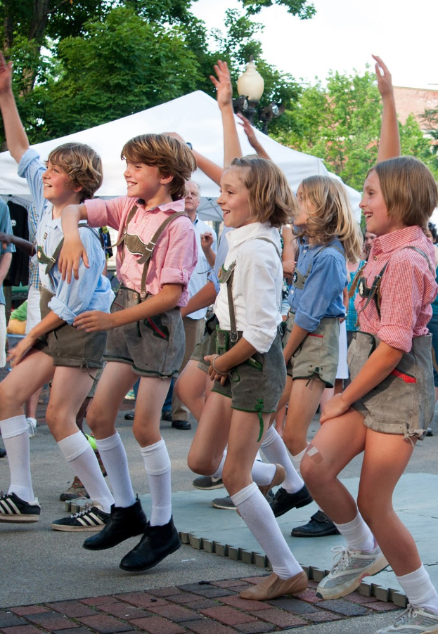 Kinder in traditioneller bayrischer Tracht, die auf einer Straße tanzen, umgeben von einer Menge, Zelten, Laternenmasten, Bäumen, Gebäuden und einem klaren blauen Himmel, wobei einige kurze Hosen und Schuhe tragen.