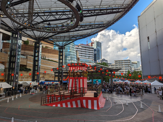 Eine große Menschenmenge umringt eine Bühne in einer städtischen Umgebung mit Gebäuden, Bäumen und einem bewölkten Himmel, wahrscheinlich während des Tokyo Circus Festivals.