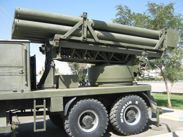 Militärlastwagen mit einer Rakete auf der Rückseite, der auf einer Straße fährt, die von Gras, Bäumen, Gebäuden und einem Maschendrahtzaun gesäumt ist.