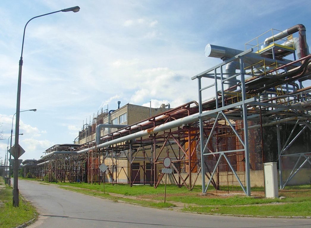 Große Fabrik mit zahlreichen Röhren an ihrer Außenseite, umgeben von Gebäuden, Straßeninfrastruktur, Vegetation und einem bewölkten Himmel.