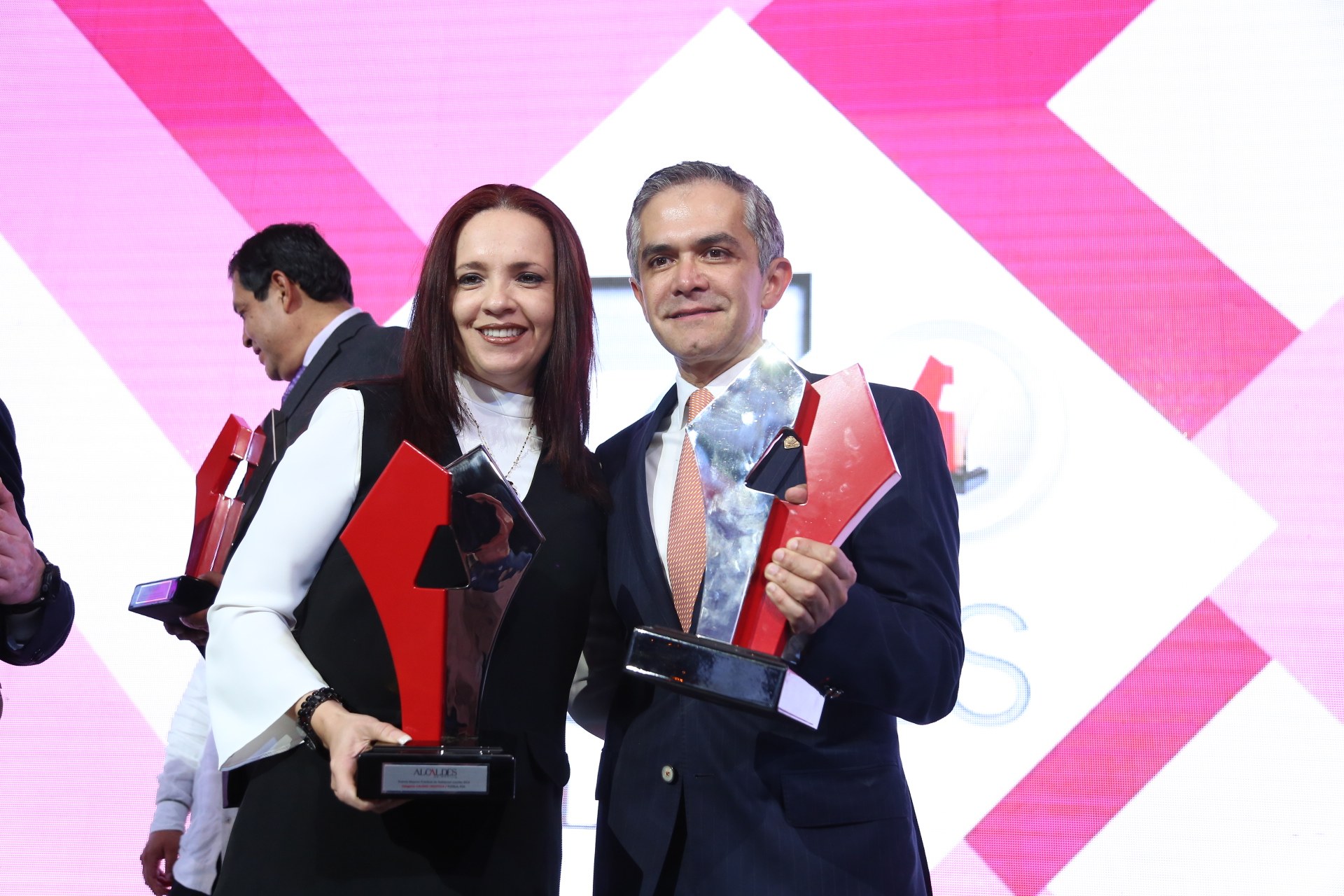 Zwei Menschen lächeln, während sie Trophäen auf einer Bühne halten.