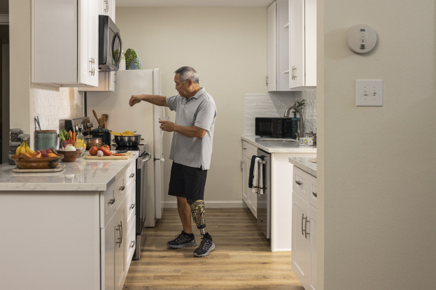 Ein Mann steht in einer Küche mit weißen Schränken, in der Hand ein Glas haltend, während verschiedene Küchenutensilien, einschließlich Obst und Besteck, auf einer Arbeitsplatte links von ihm liegen.