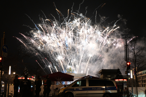 Polizeifahrzeug vor Menschenmenge bei einer Silvesterfeier in Berlin geparkt, mit farbigen Feuerwerken am Himmel.