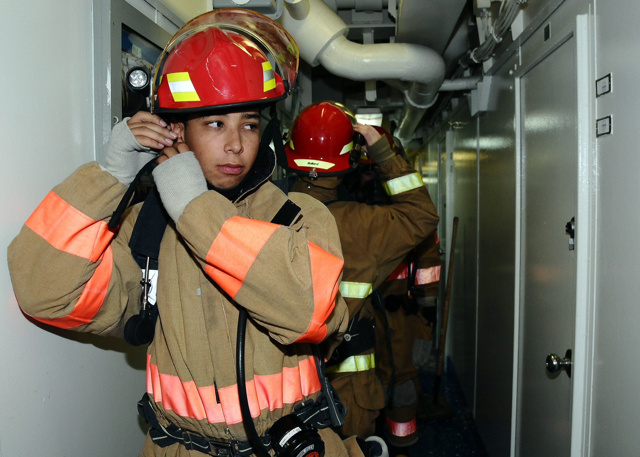 Feuerwehrleute in Uniform, die zusammen in einem Raum mit einer Tür auf der rechten Seite und Rohren im Hintergrund während einer Übung stehen.