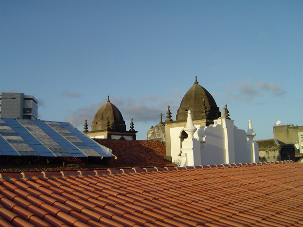 Stadtansicht mit mehreren Gebäuden im Vordergrund und einem blauen Himmel im Hintergrund, mit Solarpanelen auf einem Dach, die den Einsatz erneuerbarer Energien anzeigen.