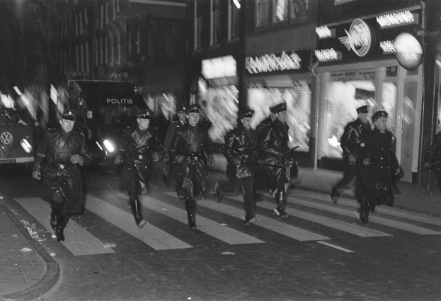 Gruppe von Polizisten in Uniform, die über eine nächtliche Straße gehen, einige mit Waffen, mit Fahrzeugen und Gebäuden im Hintergrund.