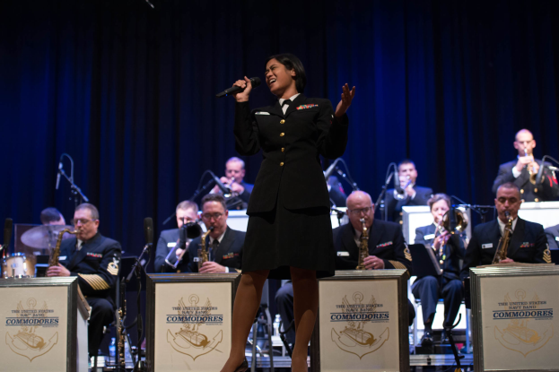 Eine Frau in militärischer Uniform steht auf einer Bühne, hält ein Mikrofon, während ein Militärorchester hinter ihr spielt.