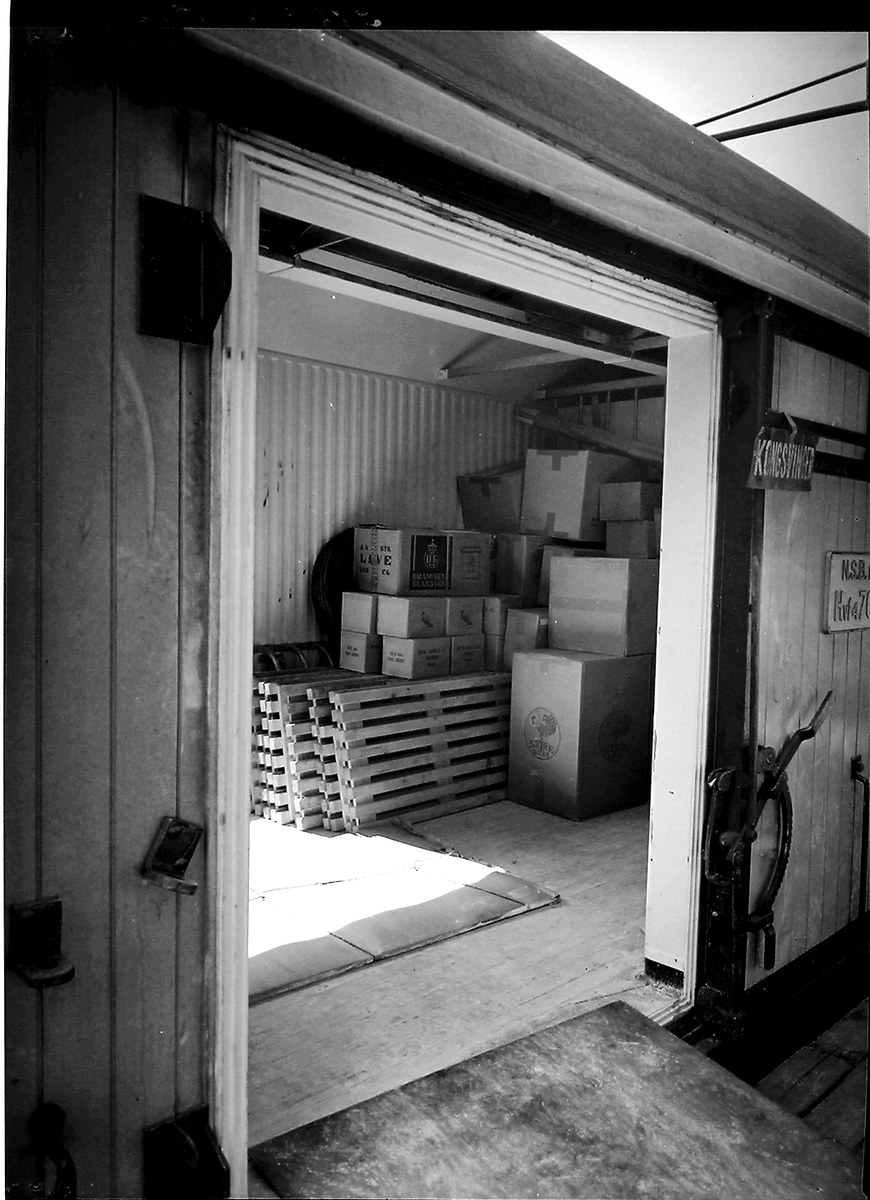 Schwarzes und weißes Foto eines Metall-Shipping-Containers mit einer Tür auf der linken Seite, gefüllt mit ordentlich gestapelten Kartons.