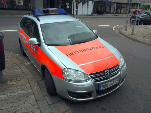 Polizeiauto am Straßenrand mit Mülltonne auf dem Gehweg zur Linken, Gebäude und Bäume im Hintergrund.